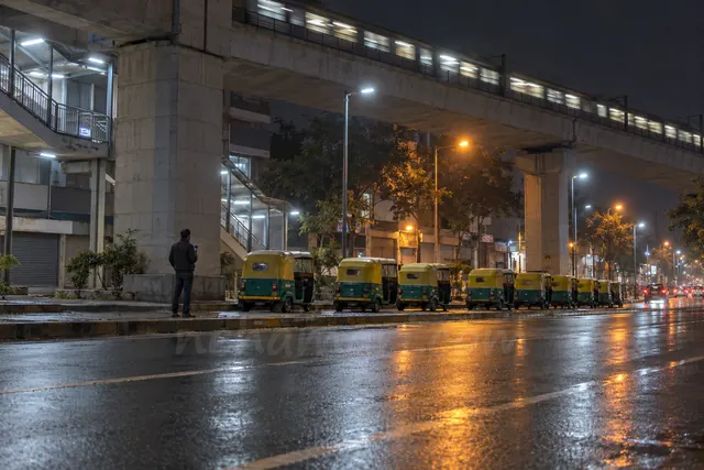 A man waits alone under the raised metro line in Bangalore at night, near a station exit, seen from across the road