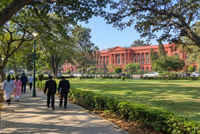 The red Karnataka High Court building in Bangalore seen from a Cubbon Park path on a weekday morning, walkers and two lawyers in black coats walking towards it