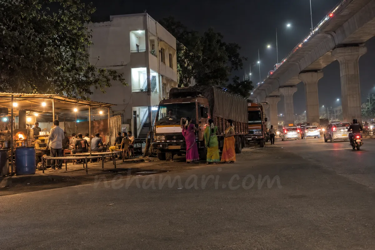 Three transgender women in bright sarees beside a parked truck, one talking to the driver, next to a roadside dhaba under the raised Electronic City expressway on Hosur Road at night
