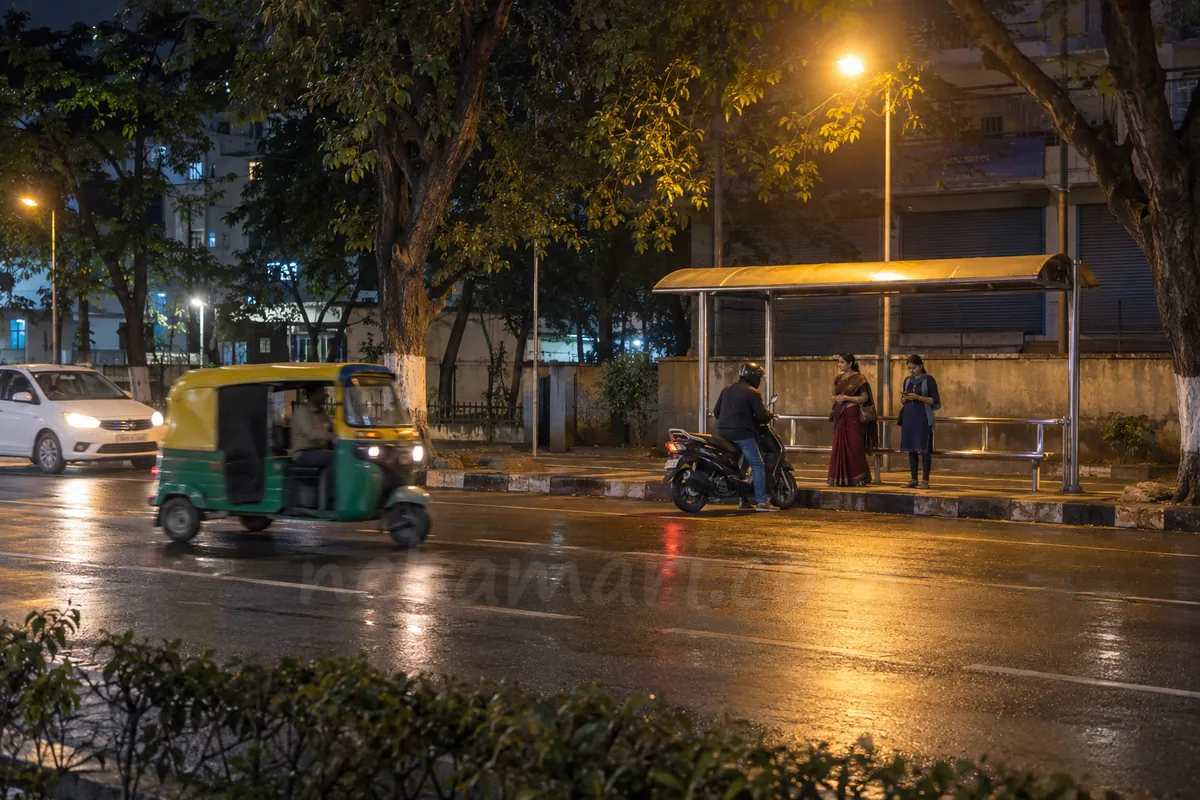 Two women wait at a steel bus shelter on a wet Bangalore road late at night while a man on a scooter stops in front of them and an auto passes