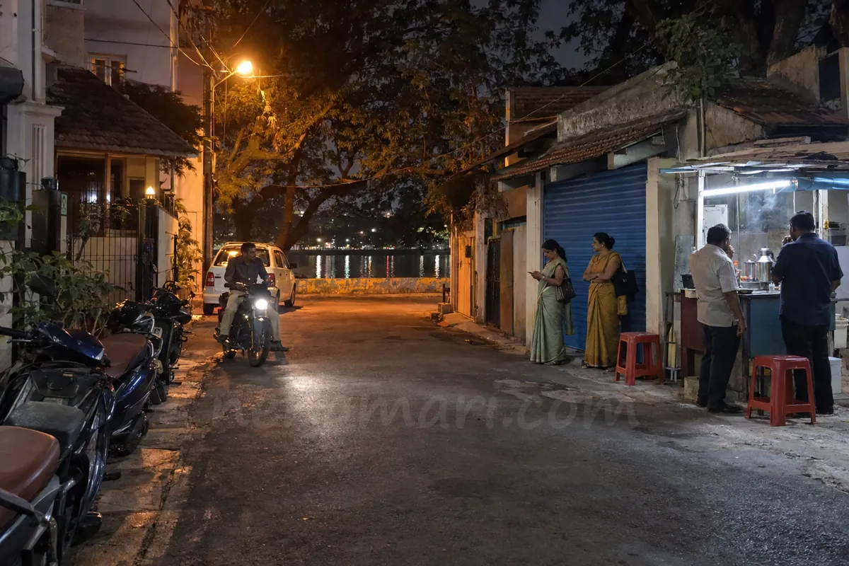 A lane in Murphy Town behind Ulsoor Lake after 10 PM: two women in sarees by a closed blue shutter, two men at a tea cart and a man on a motorbike with its light on